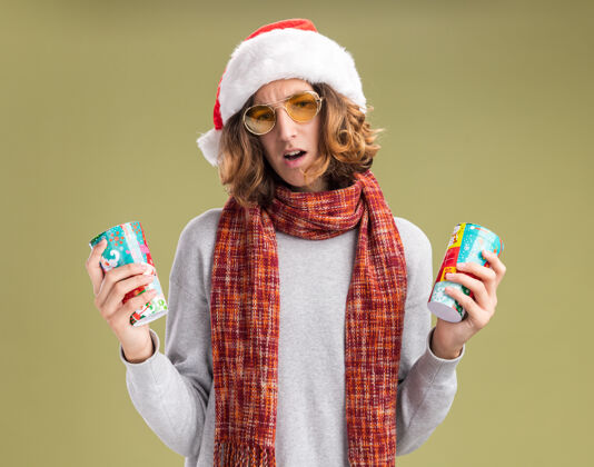 圣诞节戴着圣诞老人帽 戴着黄色眼镜 脖子上围着暖和的围巾的年轻人手里拿着五颜六色的纸杯 站在绿色的背景上 满脸疑惑眼镜围巾戴着
