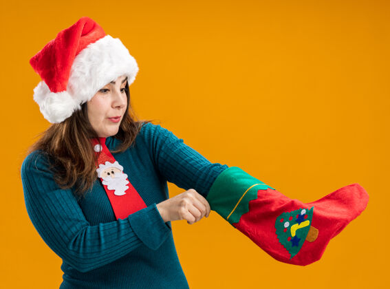 圣诞节令人印象深刻的成年白人妇女与圣诞帽和圣诞领带坚持她的手在圣诞长袜孤立的橙色背景与复制空间橙色帽子成人