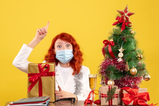 Covid19前视图女医生戴着面具坐着 圣诞礼物和黄色背景上的树病毒人女性
