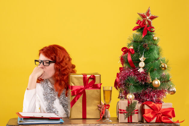 前面前视图：女医生拿着圣诞礼物和黄色背景上的圣诞树坐着背景情感快乐