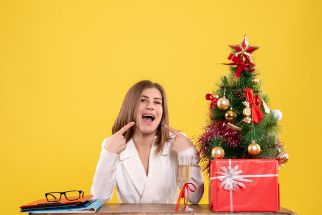 圣诞节前视图女医生坐在桌子前 带着圣诞礼物和黄色背景的树礼物树微笑