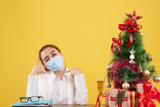 成人正面图女医生戴着防护面具坐在黄色背景上 戴着圣诞树和礼品盒女性医疗防护