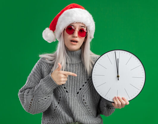 帽子年轻的金发女人穿着冬天的毛衣 戴着圣诞老人的帽子 戴着红色的眼镜 拿着挂钟 用食指指着它 站在绿色的背景下 看起来很困惑年轻指着站着