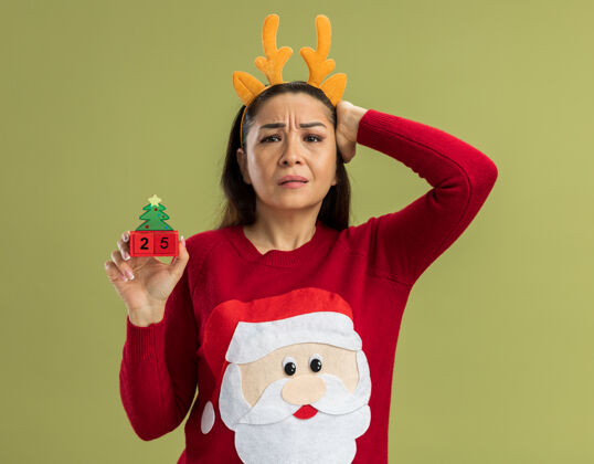 站穿着红色圣诞毛衣的年轻女子 戴着有趣的带鹿角的镶边 手里拿着玩具方块 日期是25 看起来很困惑 手放在头上 因为她忘记了站在绿色背景上困惑举行手