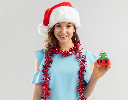高兴身穿蓝色上衣 脖子上戴着镀金圣诞帽的年轻女子 手里拿着玩具立方体 圣诞节的日子看起来很快乐帽子新的新年