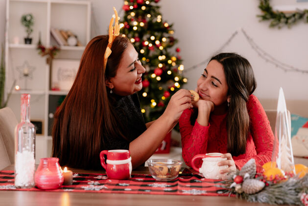 微笑微笑的母亲戴着驯鹿的头巾 喂着坐在桌边的女儿在家里享受圣诞节的快乐高兴时间圣诞快乐