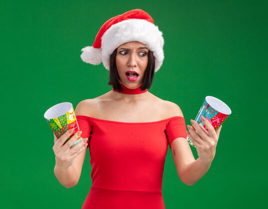杯子迷茫的年轻女孩戴着圣诞帽 手里拿着塑料圣诞杯 看着一堵孤零零的绿墙上圣诞老人帽子年轻