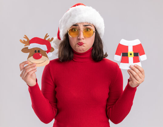 穿着穿红毛衣戴圣诞帽戴着眼镜拿着圣诞玩具困惑地站在白墙上的小女孩帽子圣诞老人年轻