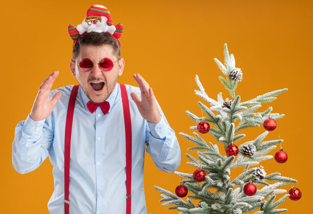 吊带戴着吊带领结的年轻人站在圣诞树旁 戴着圣诞老人和红眼镜 在橙色的墙上惊慌失措地喊叫站着男人穿着