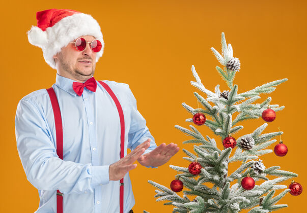 表情戴着圣诞帽 戴着红眼镜 系着吊带领结的年轻人站在圣诞树旁 手举着橘色的墙 带着厌恶的表情看着圣诞树蝴蝶结领带帽子