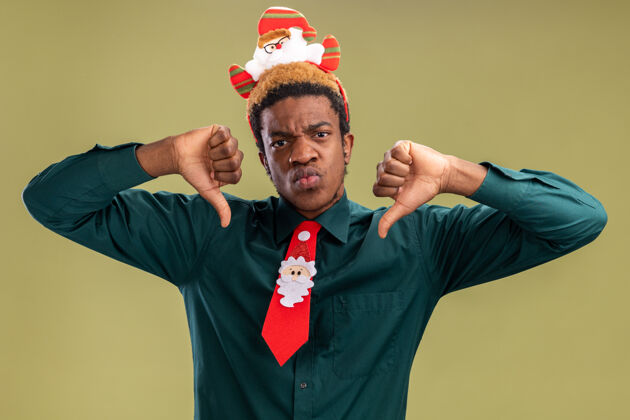 向下一个非洲裔美国人 戴着滑稽的圣诞老人戒指 打着红色领带 竖起大拇指 怒气冲冲地站在绿色的墙上拇指非洲领带