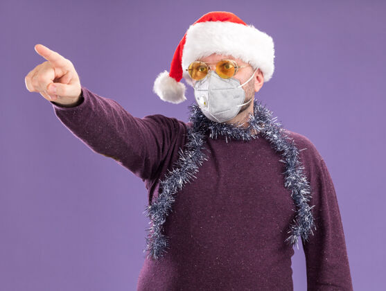 印象深刻令人印象深刻的中年男子戴着圣诞帽 戴着防护面具 脖子上戴着金属丝花环 戴着眼镜 指着隔离在紫色墙上的一侧帽子指尖圣诞老人