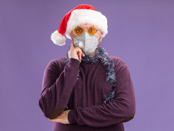 周围令人印象深刻的中年男子戴着圣诞帽 戴着防护面具 脖子上戴着金属丝花环 戴着眼镜 手放在下巴上 隔离在紫色的墙上印象深刻保护金属丝