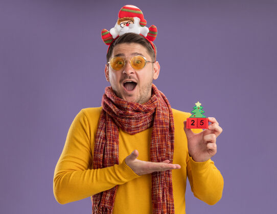 边缘快乐的年轻人穿着黄色高领毛衣 戴着暖和的围巾和眼镜 戴着滑稽的轮辋 头上戴着圣诞老人 在紫色的墙上展示着25号玩具立方体围巾穿着站立