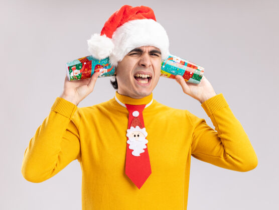表情一个穿着黄色高领毛衣 戴着圣诞老人帽 打着滑稽领带的年轻人 把五颜六色的纸杯捂在耳朵上 站在白色的墙上 带着恼怒的表情大喊大叫年轻圣诞老人站着