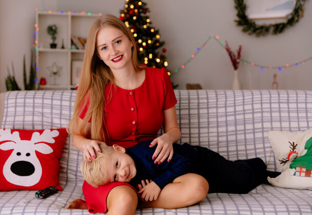 看快乐的妈妈穿着红色的裙子 带着跪在沙发上的孩子 在装饰好的房间里一起看电视 背景是圣诞树有母亲躺着