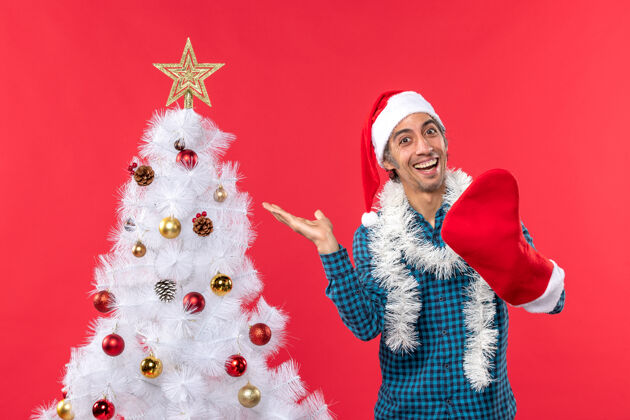 镜头一个微笑的年轻人 戴着圣诞老人的帽子 穿着一件蓝色的衬衫 穿着圣诞袜 站在红色的圣诞树旁年轻人庆祝圣诞老人