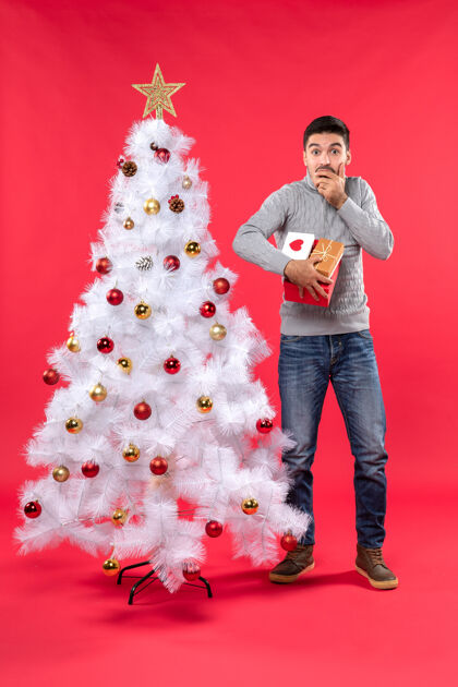 肖像迷茫的帅哥站在装饰过的白色圣诞树旁 手里拿着他的礼物礼物圣诞节成人