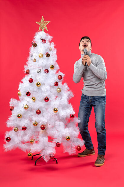 圣诞树新年气氛与震惊积极的家伙穿着牛仔裤站在装饰圣诞树附近牛仔裤站立庆祝