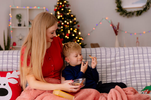 快乐在一间装饰华丽的房间里 身着红色连衣裙的快乐妈妈带着她的孩子在毯子下用饼干喂孩子 背景是圣诞树圣诞孩子圣诞快乐