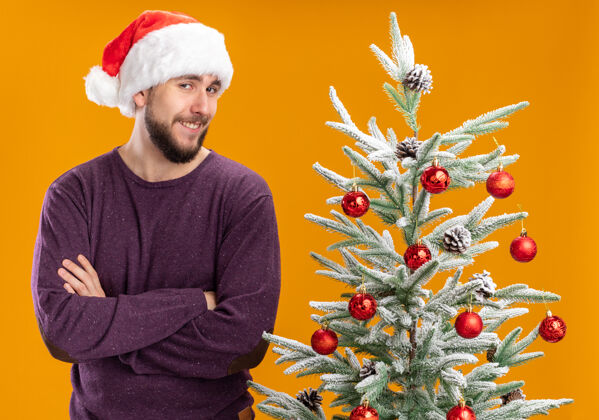 圣诞穿着紫色毛衣 戴着圣诞帽的快乐的年轻人看着相机 在橙色背景下 狡猾地微笑着站在圣诞树旁年轻人微笑圣诞树