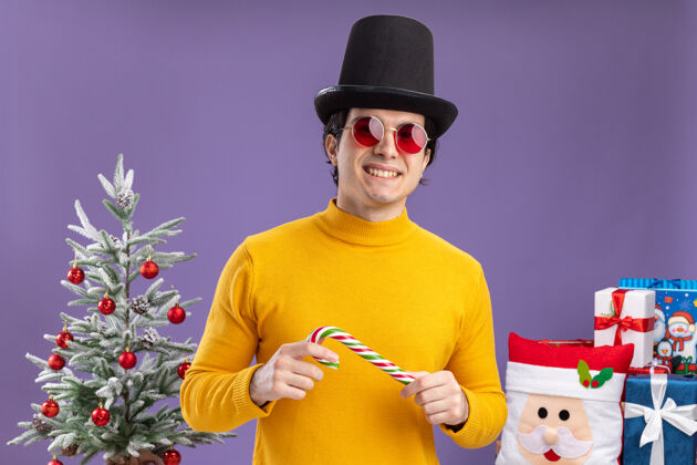 眼镜一个穿着黄色高领毛衣 戴着黑帽子 拿着糖果棒的年轻人站在一棵圣诞树旁 在紫色背景下送礼物糖果年轻人树