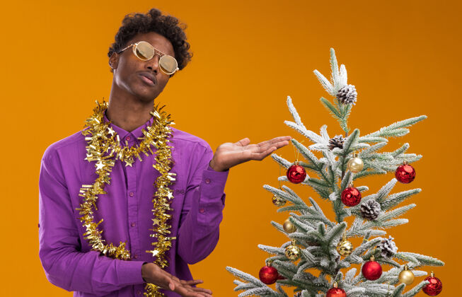 站着不知所措的年轻美国黑人男子戴着眼镜 脖子上戴着金箔花环 站在橙色背景的装饰圣诞树旁靠近圣诞不知所措