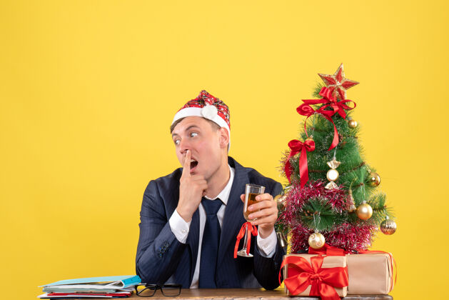 桌子困惑的商人坐在圣诞树旁的桌子前敬酒 黄色的礼物男圣诞节生意