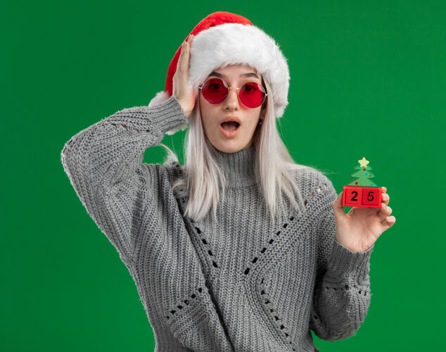 举行年轻的金发女郎穿着冬衣 戴着圣诞帽 戴着红眼镜 手里拿着玩具立方体 手里拿着圣诞节的日期 站在绿色的背景下 看着相机 惊讶又惊讶站着惊讶金发