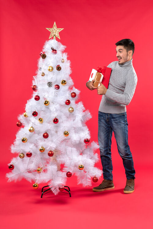 礼物帅气的年轻人站在装饰好的白色新年树旁 手里拿着礼物衣服圣诞树