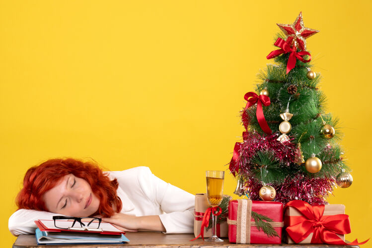 办公室女工人拿着圣诞礼物坐在桌子后面 树睡在黄色的地板上礼物黄色女