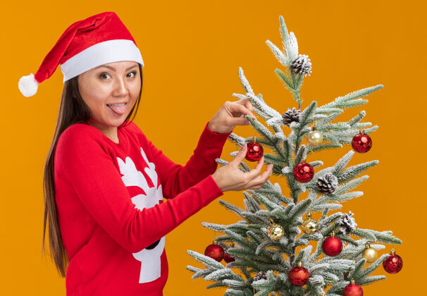 舌头快乐的年轻亚洲女孩戴着圣诞帽和毛衣装饰圣诞树 在橙色的墙上露出孤立的舌头装饰圣诞节年轻