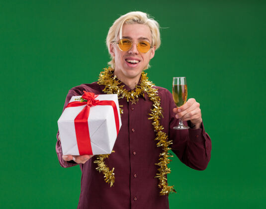 年轻快乐的年轻金发男子戴着眼镜 脖子上戴着金箔花环 手里拿着一杯香槟 把礼包摊开放在绿色的墙上圣诞节金属丝快乐