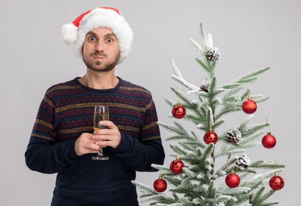 气喘吁吁令人印象深刻的年轻白种人戴着圣诞帽站在圣诞树旁 举着一杯香槟 在白色的墙壁上孤立着鼓鼓的脸颊年轻站着戴着