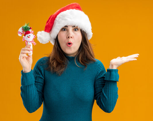 圣诞老人兴奋的成年白人妇女戴着圣诞帽 手持糖果手杖 在橙色背景上保持手张开 并留有复制空间背景保持圣诞快乐
