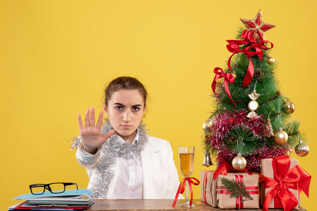 坐着正面图：女医生坐在黄色背景的桌子后面 还有圣诞树和礼品盒女情感插花