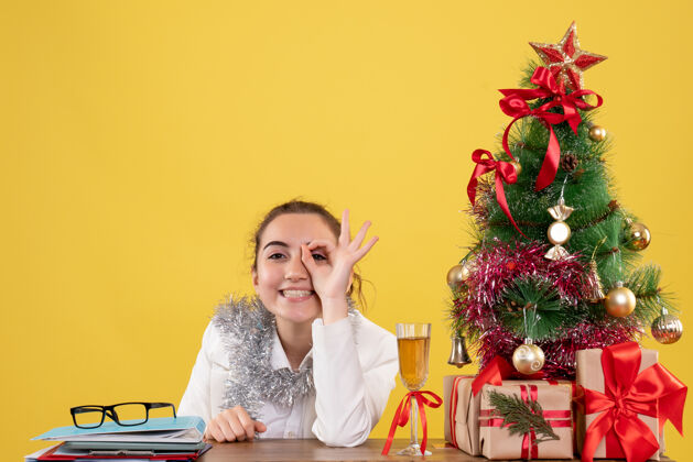 前面正面图：女医生坐在桌子后面 在黄色背景下微笑着 手里拿着圣诞树和礼品盒健康插花圣诞节
