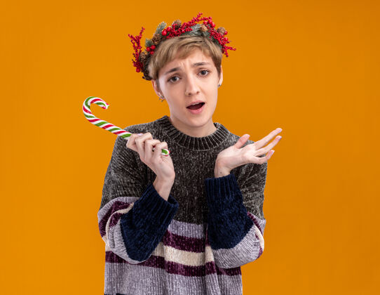年轻笨手笨脚的年轻漂亮女孩戴着圣诞花环 手持圣诞甜手杖 手上空空的 被隔离在橙色的墙上 留着复制空间头花环穿着