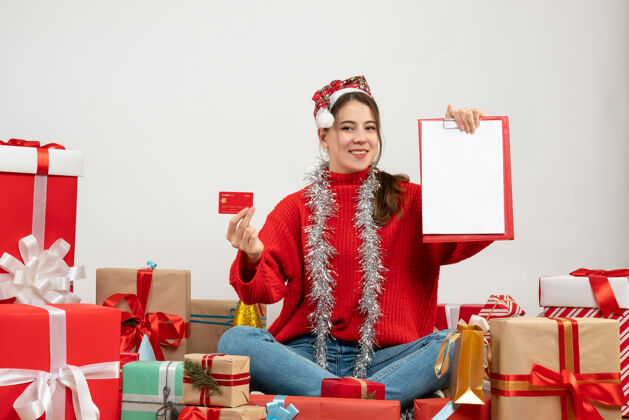 文件带着圣诞帽的快乐派对女孩拿着卡片和文件坐在白色的礼物旁边周围女孩圣诞老人