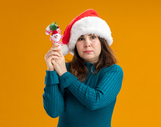 橘子焦虑的成年白人妇女 戴着圣诞帽 手里拿着糖果手杖 被隔离在橙色背景上 还有复制空间圣诞老人手杖帽子