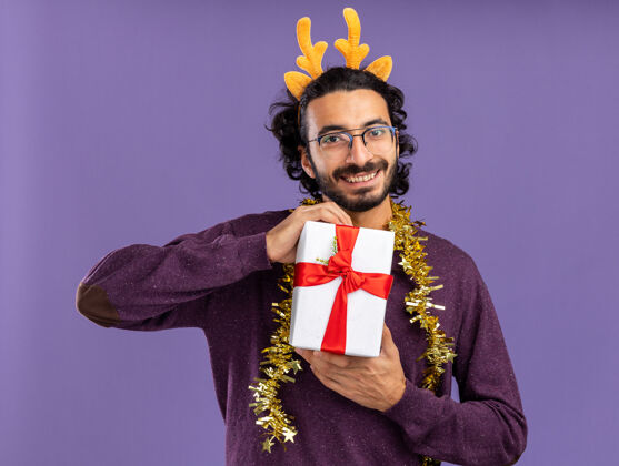 微笑微笑的年轻帅哥戴着圣诞发箍 脖子上戴着花环 手里拿着蓝色背景的礼品盒礼物年轻花环