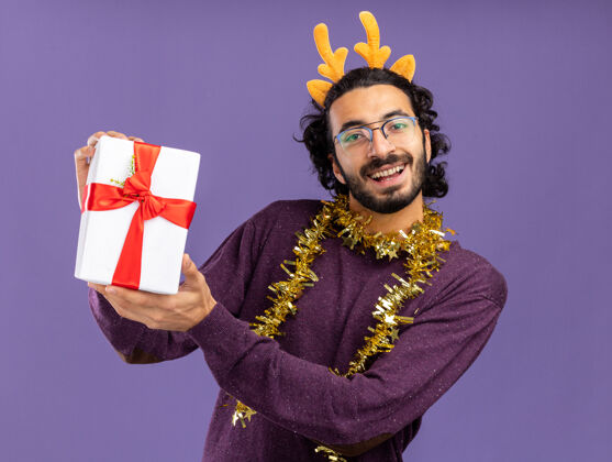花环微笑的年轻帅哥戴着圣诞发箍 脖子上戴着花环 手里拿着蓝色背景的礼品盒小伙子脖子箍