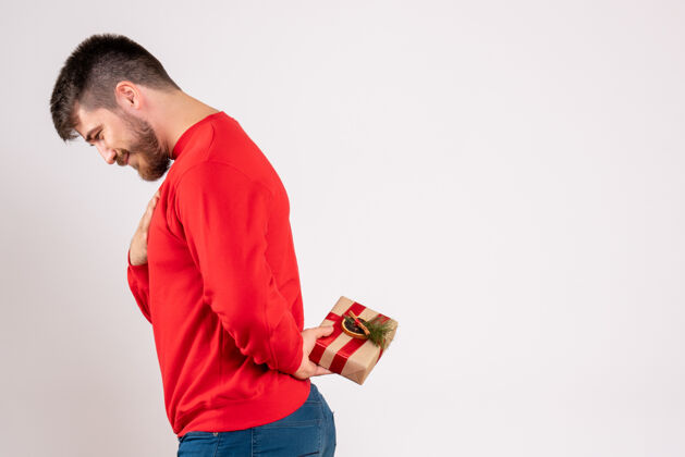 节日一个穿着红衬衫的年轻人把圣诞礼物藏在他背后的白墙上年轻礼物背