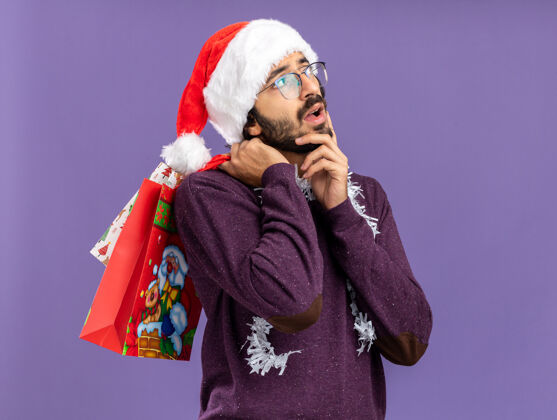 肩膀年轻帅哥戴着圣诞帽 脖子上戴着花环 肩上挎着礼品袋 手放在脸颊上 蓝色的背景上与世隔绝脸颊花环手