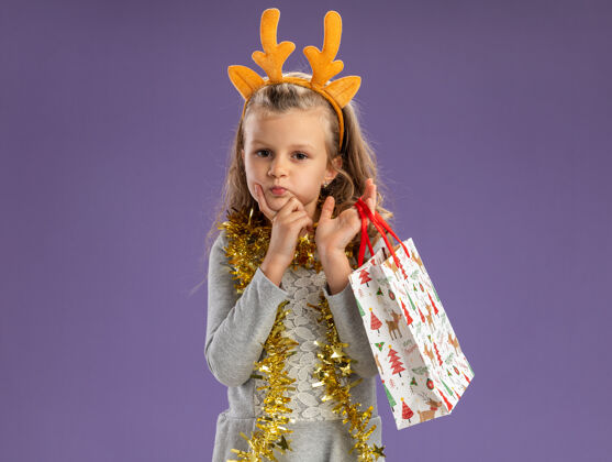 把想着小女孩戴着圣诞发箍 脖子上戴着花环 手里拿着礼品袋 手放在下巴下面 蓝色背景上孤立着花环小包