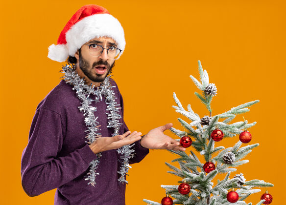 站着困惑的年轻帅哥站在圣诞树旁 戴着圣诞帽 脖子上戴着花环 指着隔离在橘色墙上的圣诞树点困惑圣诞节