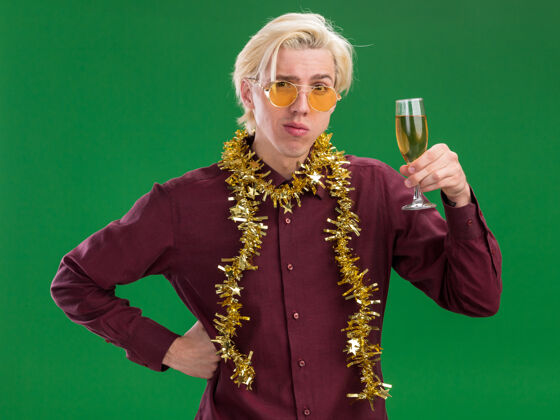 年轻可疑的年轻金发男子戴着眼镜 脖子上戴着金箔花环 手里拿着香槟酒杯 手放在腰上 隔离在绿色的墙上圣诞节男人花环