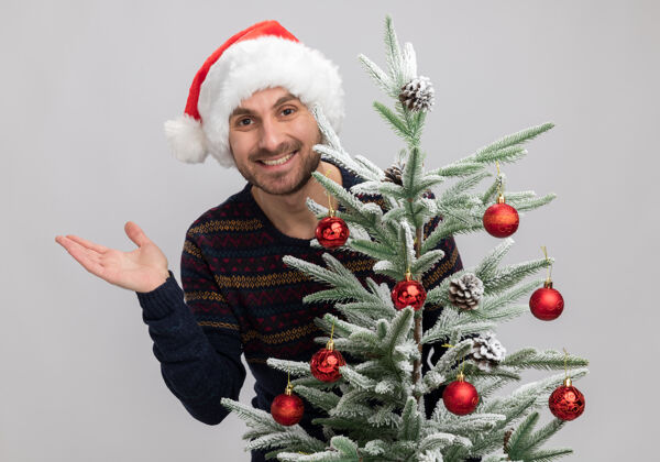 帽子快乐的白人年轻人戴着圣诞帽站在圣诞树后 在白色的墙上孤零零地伸出空手戴着站着男人