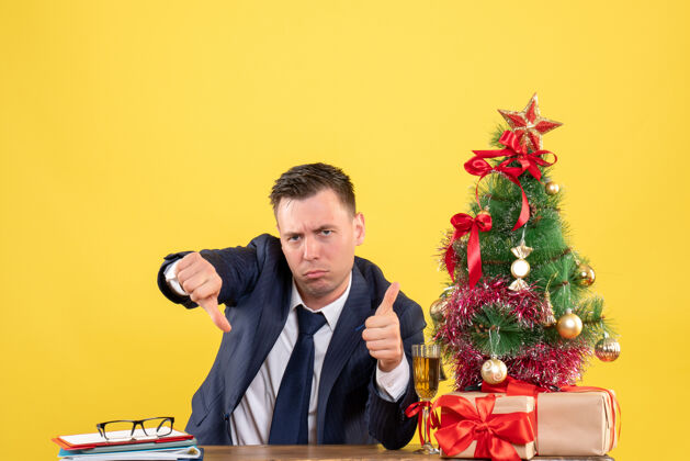 制造前视图严肃的男子 使拇指上下标志坐在圣诞树和黄色礼物附近的桌子上圣诞节男人坐着