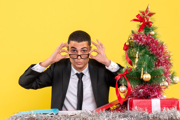 男人前视图一个穿着西装的年轻人坐在桌子旁摘下眼镜圣诞树和礼物桌子圣诞节拿着